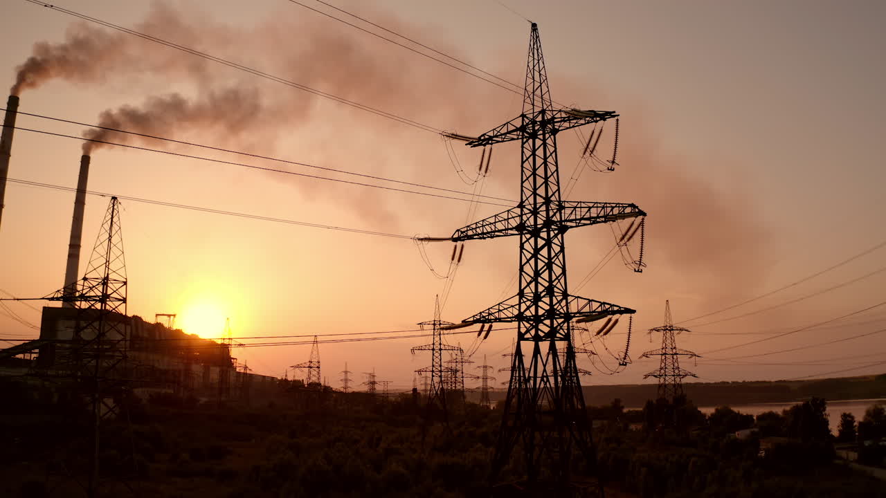 Power station at dusk. Transmission lines near smoke pipes from industrial plant. Chemical emissions in the air from industry. Electric towers with wires at sunset.