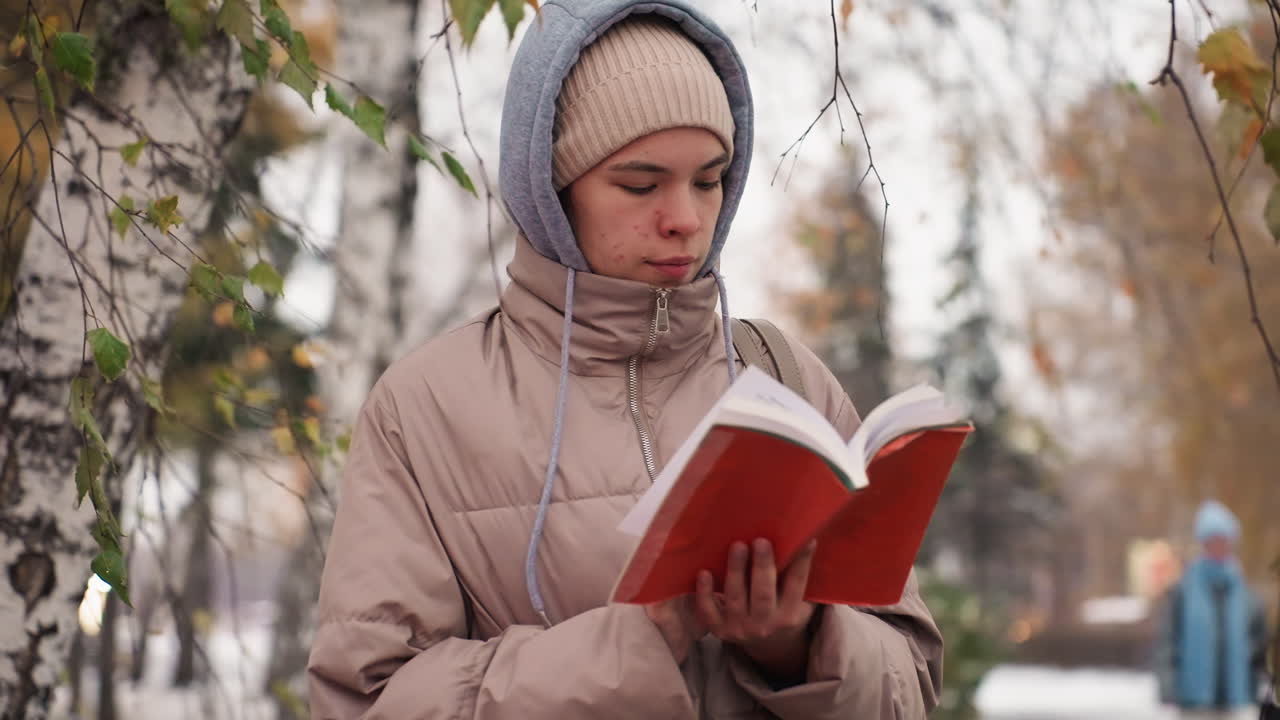 Light skin lady wearing winter jacket and knit hat reading red book, flipping page while standing close to tree in serene autumn setting with softly blurred background and falling leaves