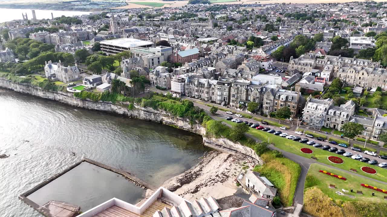 Drone camera glides above St Andrews’ historic coastline, revealing tidal pools, stone buildings, and green spaces under bright, diffuse daylight