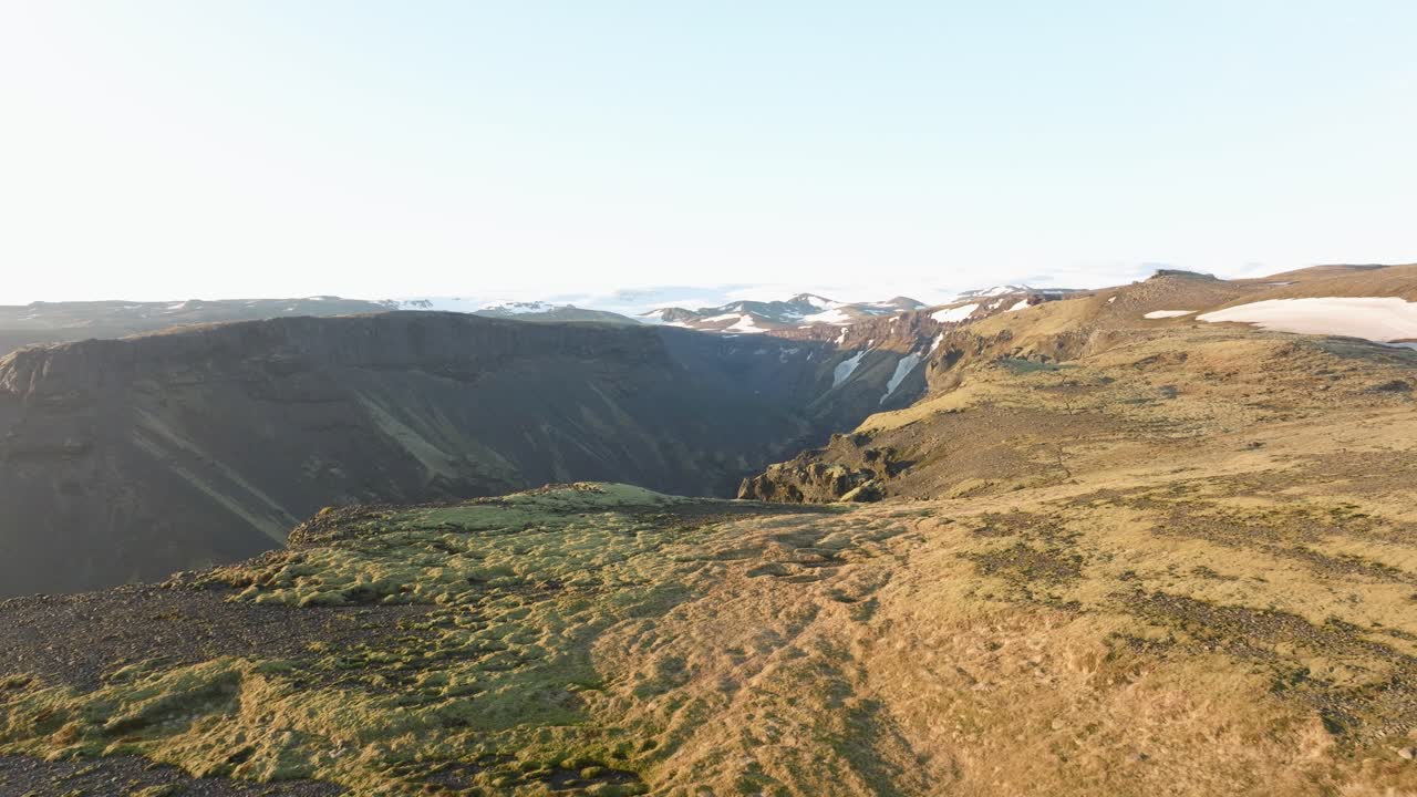 Drone shot of Klfurárgil canyon in Iceland, featuring rugged cliffs, green highlands, and snow-capped mountains under clear skies