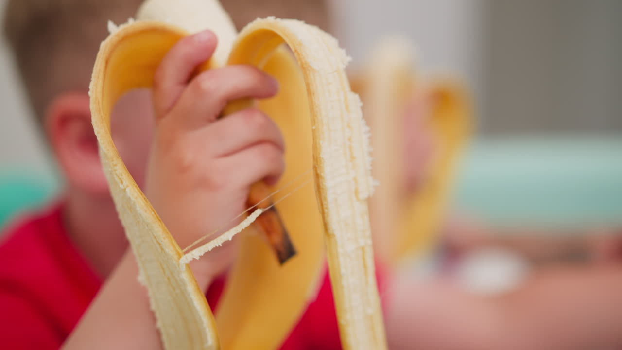 Close up of young boy eating banana while sitting in pastel ball pit, playing and enjoying snack time with blurred view of another child also eating banana and playing in cheerful indoor environment