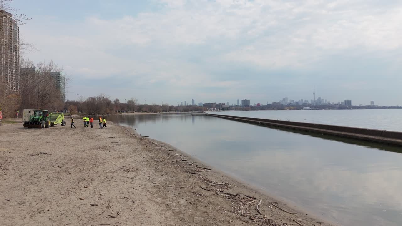 Aerial: city workers on the beach next to Lake Ontario with downtown Toronto skyline and CN Tower in the back, jib down drone shot