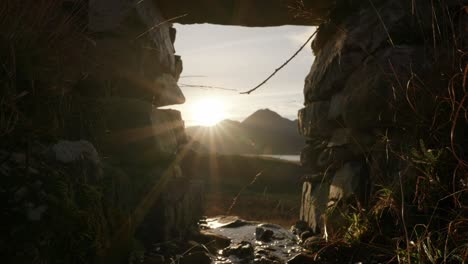 the sun sets behind a scottish mountain producing a sunburst in the background as a small river gently flows under an arch in a stone wall