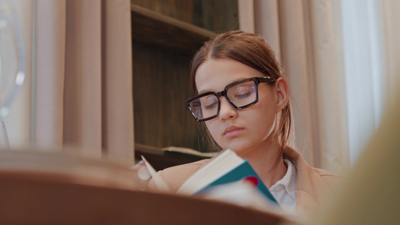 Primer plano de mujer leyendo un libro en el estudio, luz natural suave, gafas negras y cabello trenzado, blazer neutro, escritorio de madera y estantería de fondo, concentración tranquila y paso de página, estado de ánimo reflexivo y sereno.