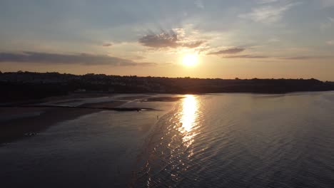 Sunset-rays-beam-fiery-golden-colours-across-relaxing-beach-ocean-coastline-aerial-view-descend-to-above-water