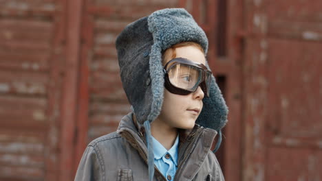 close up of a cute little red haired boy in aviator hat and glasses turning face and smiling cheerfully at the camera