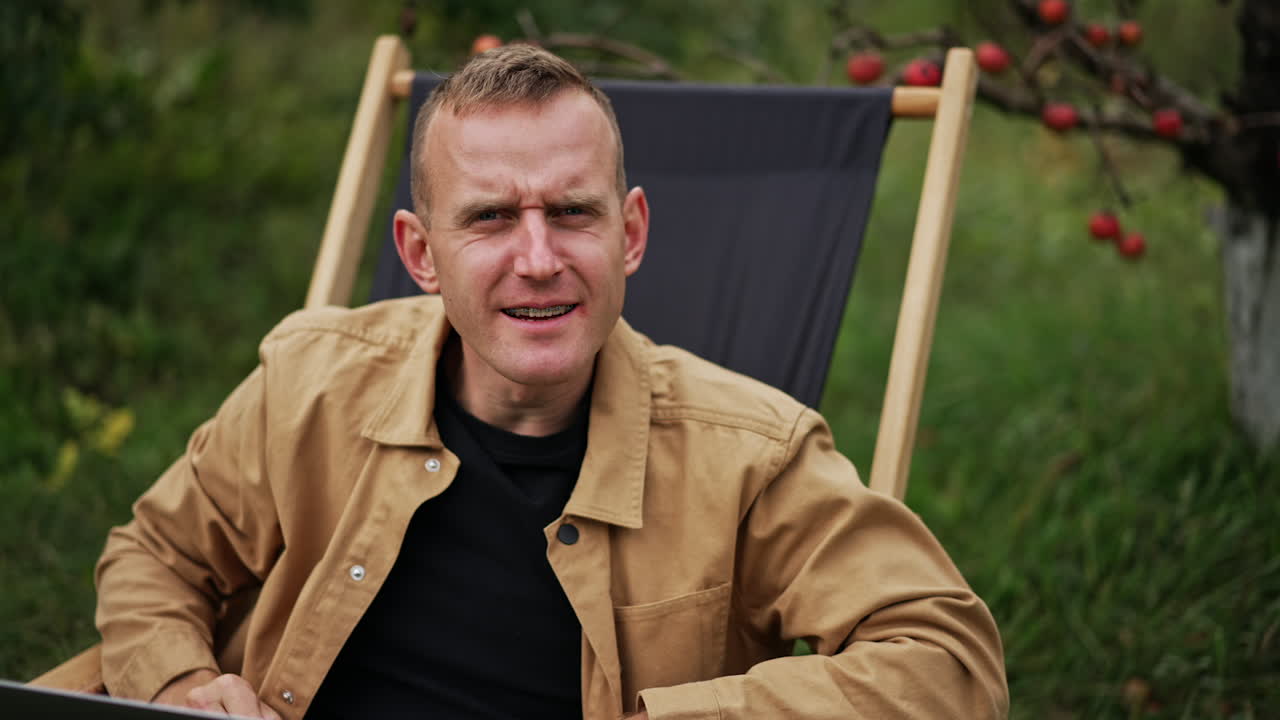 Freelance worker man in a chair looks strictly into camera. Male working remotely in his garden smiling to the camera on his laptop. Blurred nature backdrop.