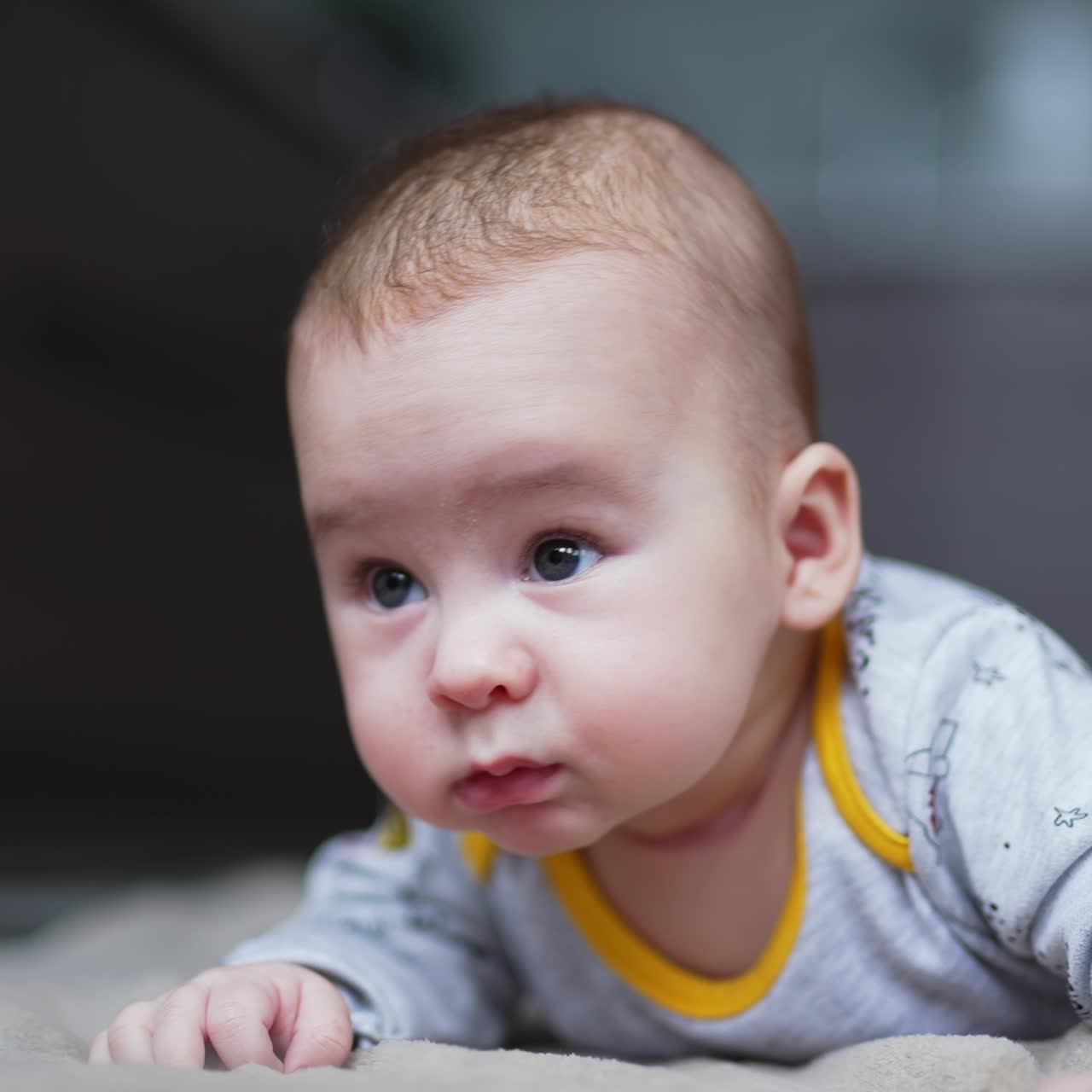 Nice cute boy lies on stomach and keeps head up. Beautiful child with adorable plump cheeks on grey blurred backdrop
