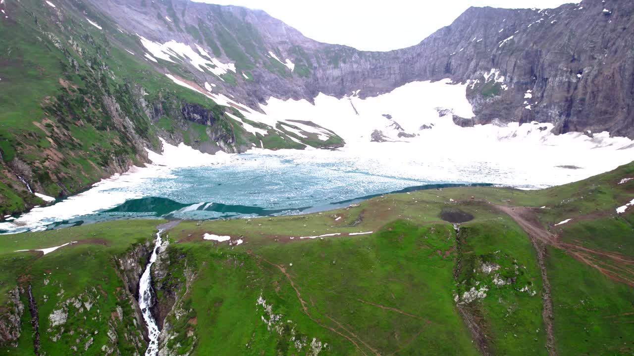 lago de montaña rodeado de picos cubiertos de nieve, con una cascada que fluye por un acantilado empinado