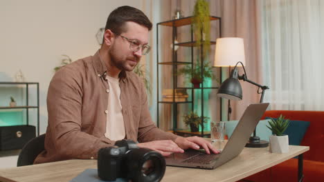 young caucasian man photographer transfer data files from camera to laptop at home office table desk