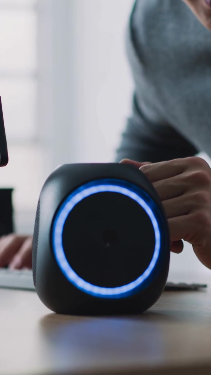 Video verticale: Uomo con maglietta grigia che nota un LED blu luminoso, appoggiato alla scrivania mentre parla a uno smart speaker.