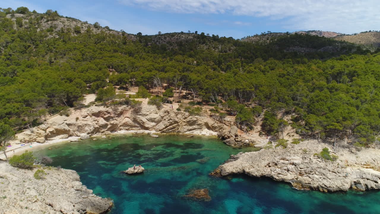 tomada aérea de un avión no tripulado de una bahía con agua cristalina y rocas y árboles en un día soleado, mallorca, españa, volando hacia adelante, 4k