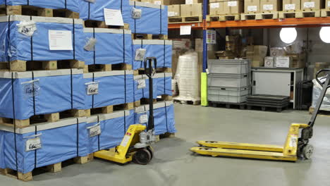 Un-Trabajador-De-Almacén-Con-Chaleco-Reflectante-Camina-Entre-Filas-De-Palés-Envueltos-En-Azul,-Utilizando-Una-Transpaleta-Manual-Con-Rodillos-Dobles.