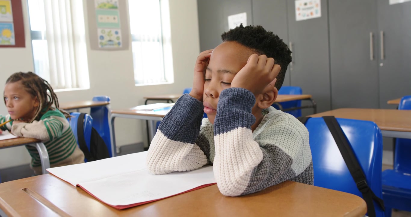 Compagni di classe afroamericani che guardano un ragazzo al banco in classe perdere la concentrazione e appoggiare la testa sul quaderno.