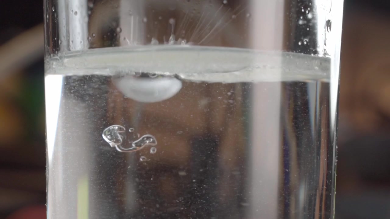 Primo piano macro di gocce di liquido che cadono in un liquido trasparente all'interno di un bicchiere, creando piccole bolle d'aria e increspature sotto la superficie con goccioline visibili sul vetro.