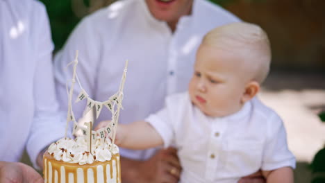 A birthday cake for one year of life is held in front of a little blond baby. Kid in dad's hands trying to reach a dessert.