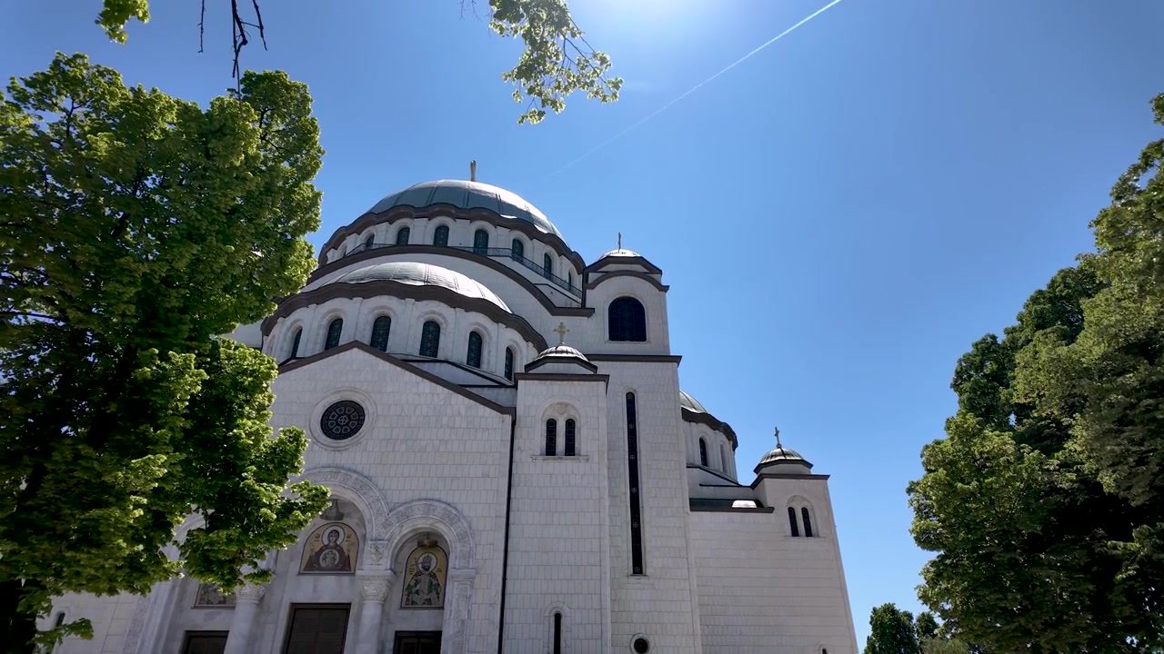 Uma igreja é visível sob um céu azul claro. Árvores altas cercam a área, e uma estátua fica nas proximidades. O cenário mostra um dia ensolarado com um leve rasto de nuvem de um avião.