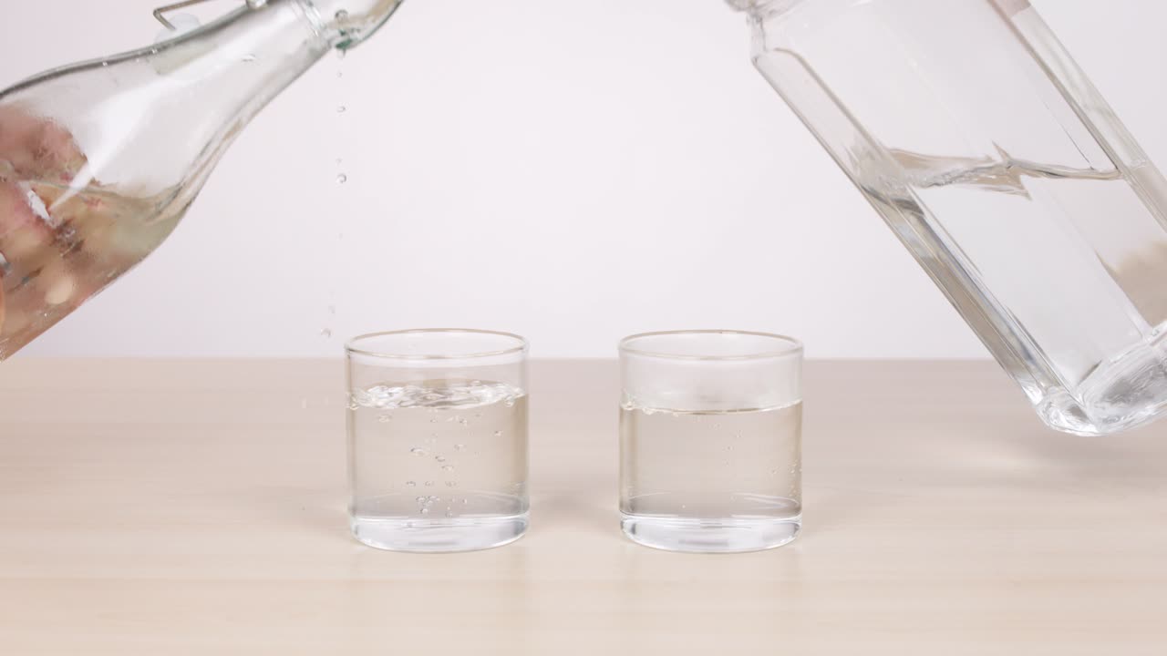 Two glass bottles pour water into separate glasses on a light table, bright studio lighting