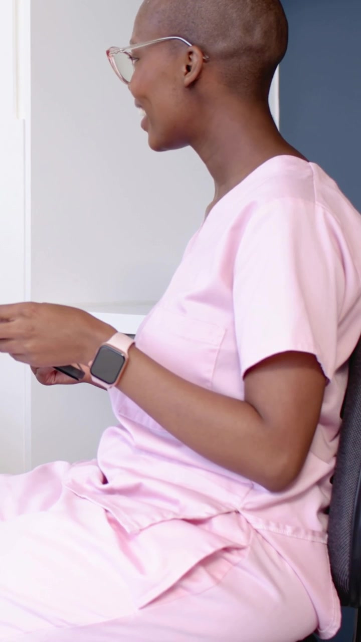 Video verticale: Donna in divisa medica rosa che tiene in mano il telefono, parla e gesticola in una clinica, indossando uno smartwatch.