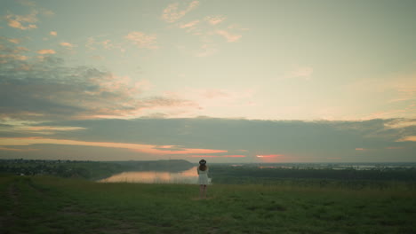 solitary woman in white dress and hat stands in a peaceful grassy field, overlooking a tranquil lake and distant village under a pastel-colored, cloud-streaked sky at sunset