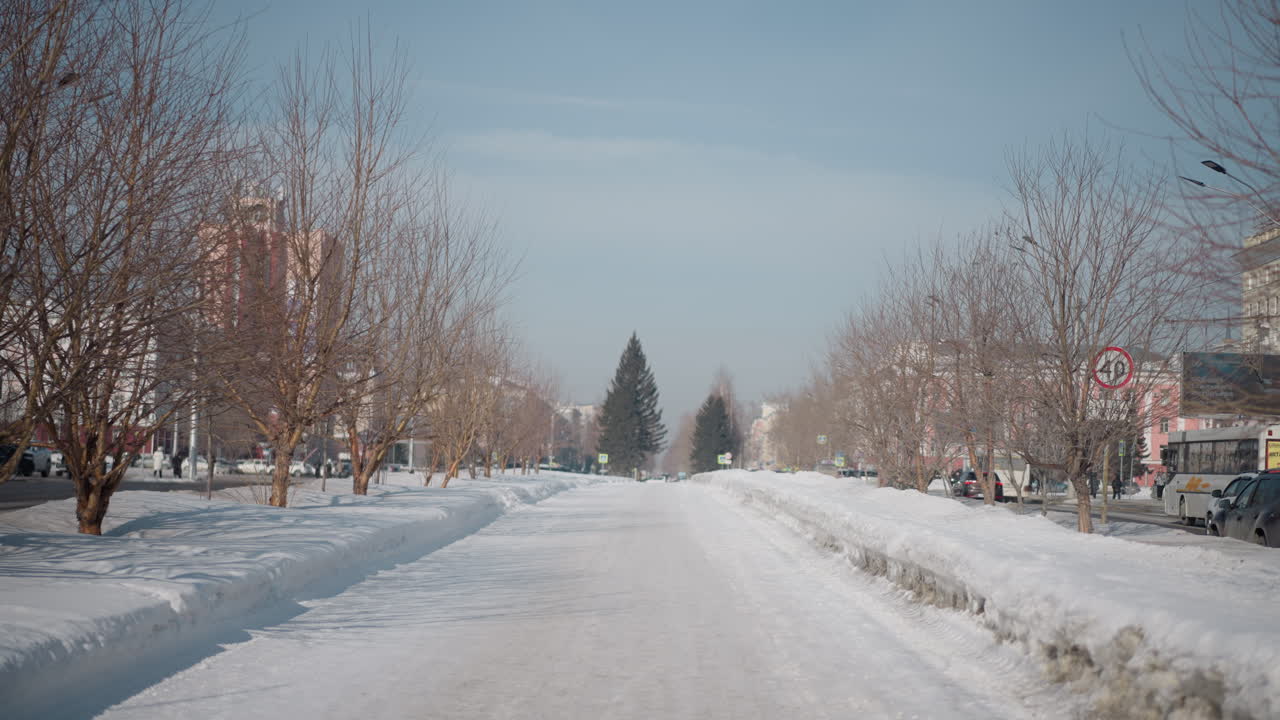 long view along snowy median road bordered by leafless trees, cars pass on both sides under pale blue sky, plowed banks line route, cold winter light and distant perspective create calm urban commute