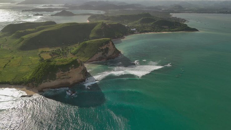 Filmato aereo con drone della baia per surf di Gerupuk con onde che si infrangono sulla barriera corallina, linee di surf bianche, acqua turchese, scogliere sul mare, colline verdi e piccole barche al largo, paesaggio costiero cinematografico in calda luce serale 4k.