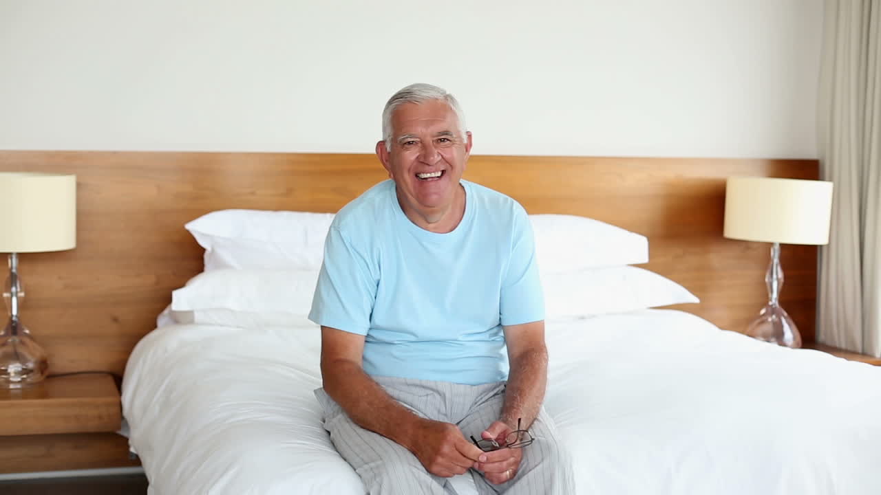 Uomo anziano seduto sul letto che sorride alla telecamera al mattino