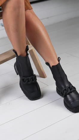 lady model sitting on a wooden stool showing the fashionable stylish footwear. black leather boots with chain décor on a female legs. vertical video
