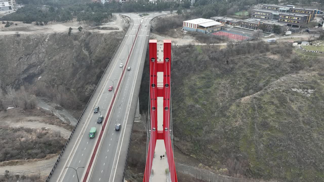 A long red bridge spans across a rugged winter gorge, connecting urban and rural elements with its modern design. Cars traverse the structure, adding a dynamic touch to the serene scene