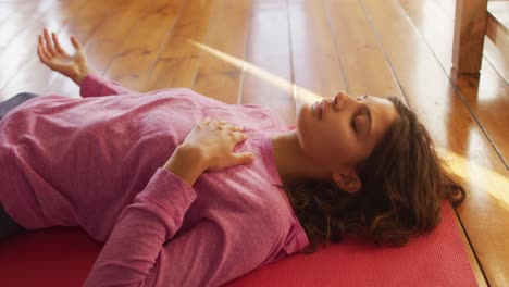 Relaxed-mixed-race-woman-practicing-yoga,-lying-on-mat-with-eyes-closed-in-sunny-cottage-bedroom