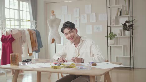young asian male fashion designer drawing pencil sketches of clothing and smiling to the camera