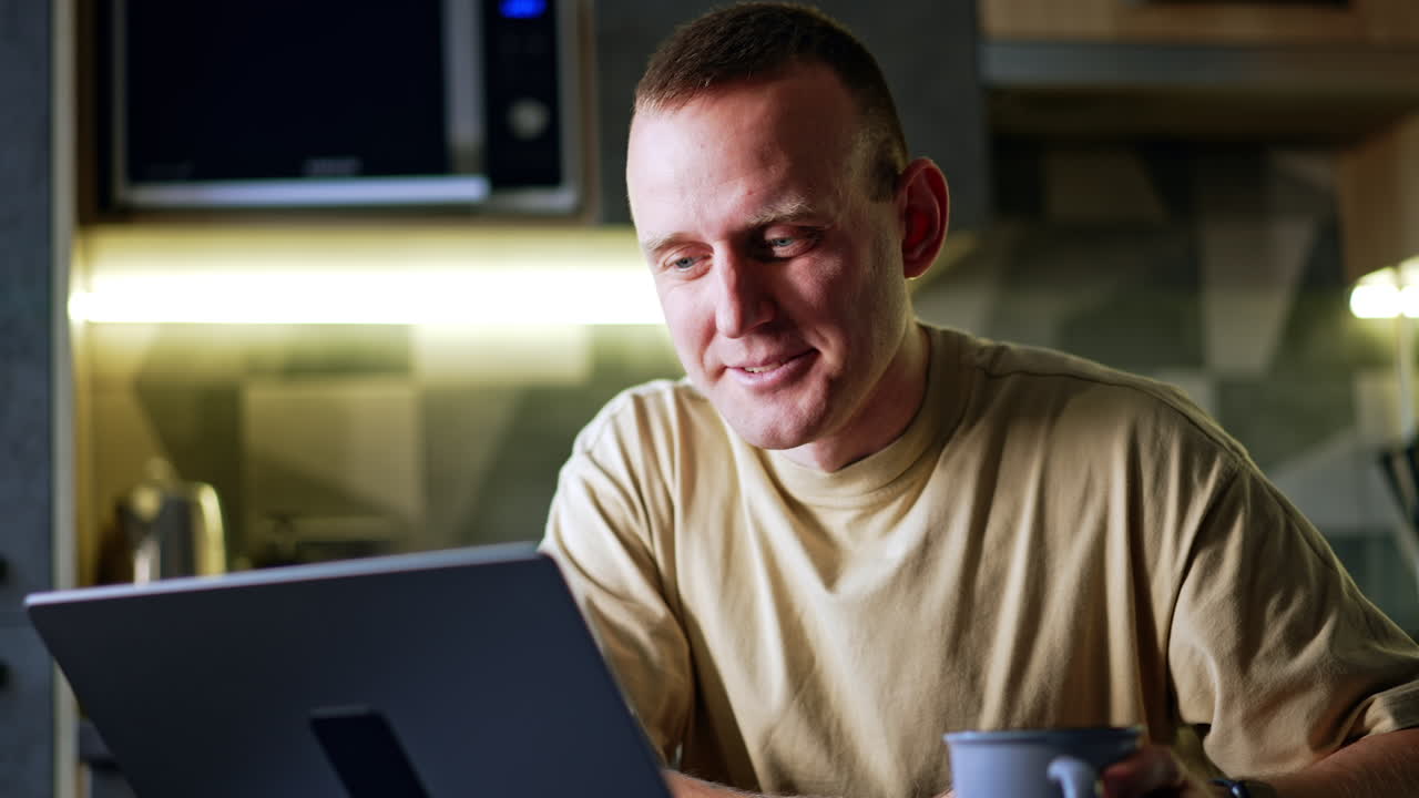 Smiling Caucasian male sits looking at the screen of his laptop. Man works in his kitchen remote from office. Close up portrait.