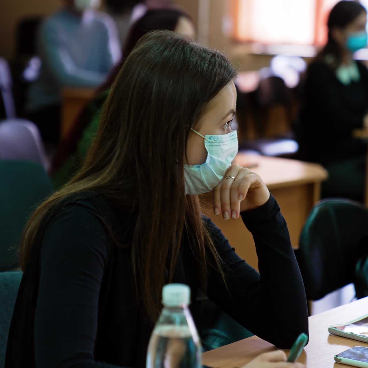 Long-haired brunette woman wearing mask sitting at the desk. Woman taking notes listening to the lecturer. Lots of students at backdrop