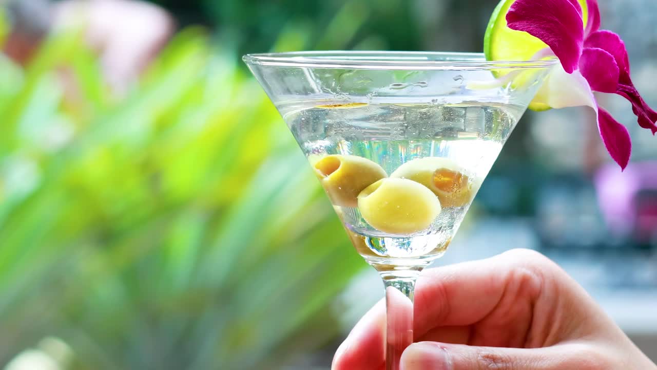 A close-up of a martini glass with olives and an orchid garnish, held against a blurred green background.