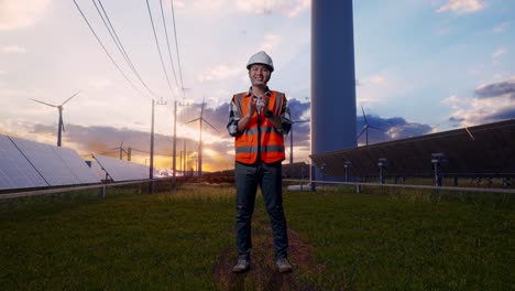 full body of asian male engineer with safety helmet smiling and clapping his hands while standing with solar panel and wind turbines
