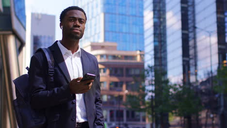 Young-Businessman-Wearing-Wireless-Earbuds-Using-Mobile-Phone-App-To-Hail-Taxi-Cab-In-The-Financial-District-Of-The-City-Of-London-UK