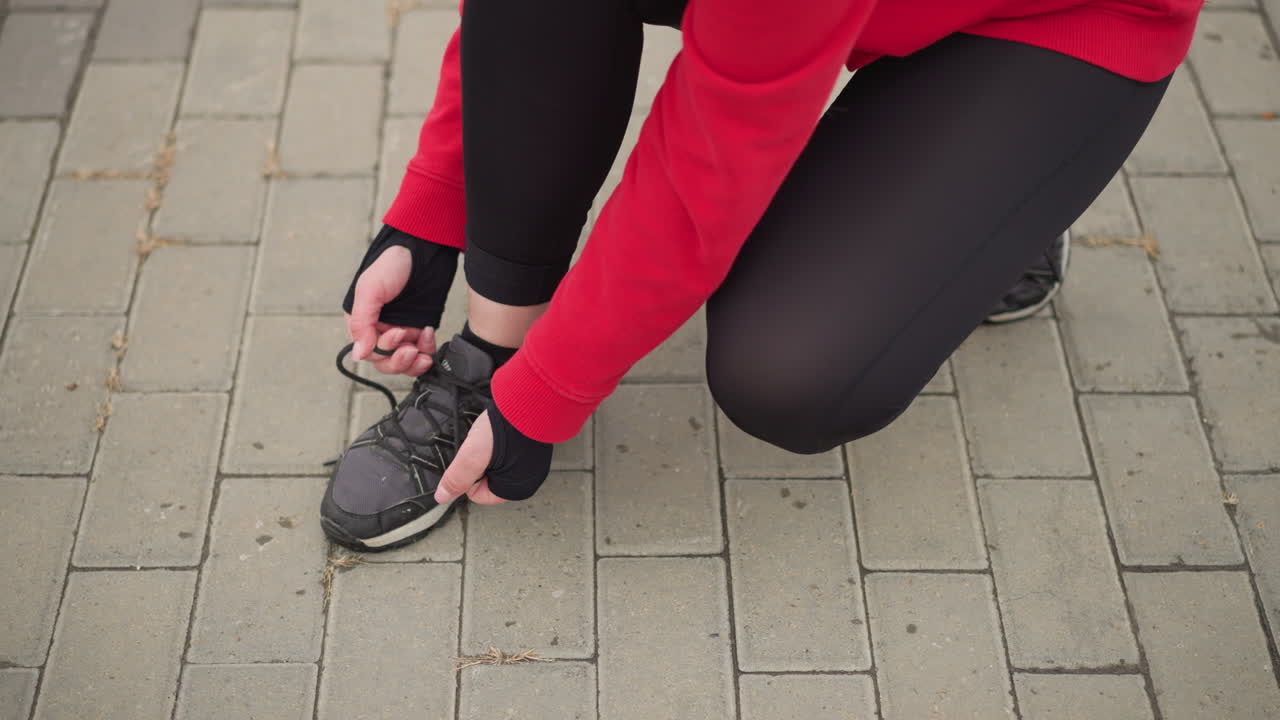 Side view of athlete squatting on interlocked path tying her right shoelace, close-up focus on hands and shoe, highlighting preparation and dedication for outdoor activity in fitness