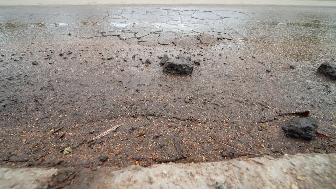 Primer plano en ángulo bajo de una rueda de coche pasando sobre asfalto mojado y agrietado con baches y escombros invernales, mostrando las difíciles condiciones de la carretera después de la temporada de deshielo y congelación.