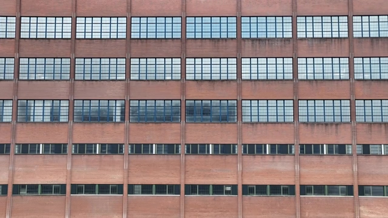 Una vista frontal de un alto edificio de oficinas de ladrillo ocupa la mayor parte del encuadre, con la fachada principal centrada y una extensión más estrecha visible en el borde derecho.