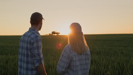 una pareja de agricultores jóvenes miran cómo trabaja un tractor en el campo. de pie uno al lado del otro contra el telón de fondo de un campo de trigo donde se está poniendo el sol