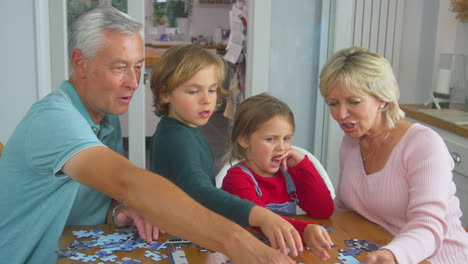 Los-Nietos-Con-Los-Abuelos-Se-Sientan-Alrededor-De-La-Mesa-En-Casa-Haciendo-Rompecabezas-Juntos.