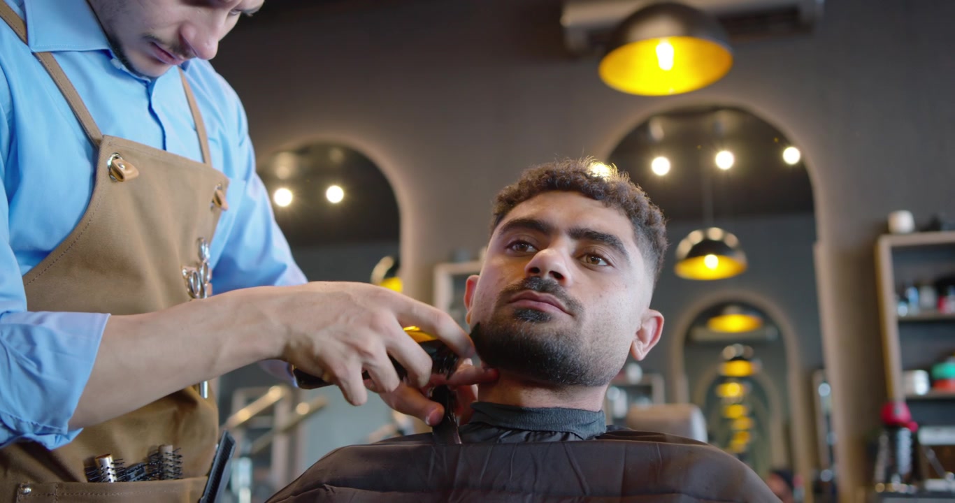 Barber kommer fra venstre, former skæg med trimmer på kunde iført sort kappe i barbersalon
