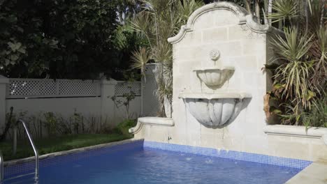 Poolside-fountain,-water-trickles-down-into-pool-before-vegetation-and-greenery-in-background