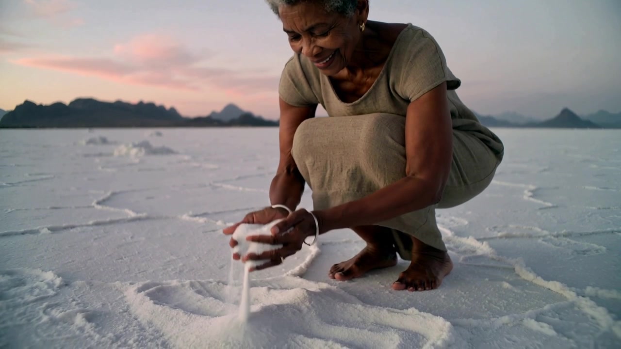 Kvinde i bevægelse danner en bunke, skovler og hælder salt på en saltslette ved solnedgang, iført olivenfarvet kjole.
