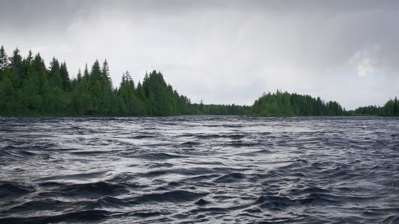 Wide view of Glomma river in Norway filmed at dusk, with dark rippling water and dense coniferous forest under an overcast sky, conveying a moody and tranquil Nordic atmosphere in slow-motion.