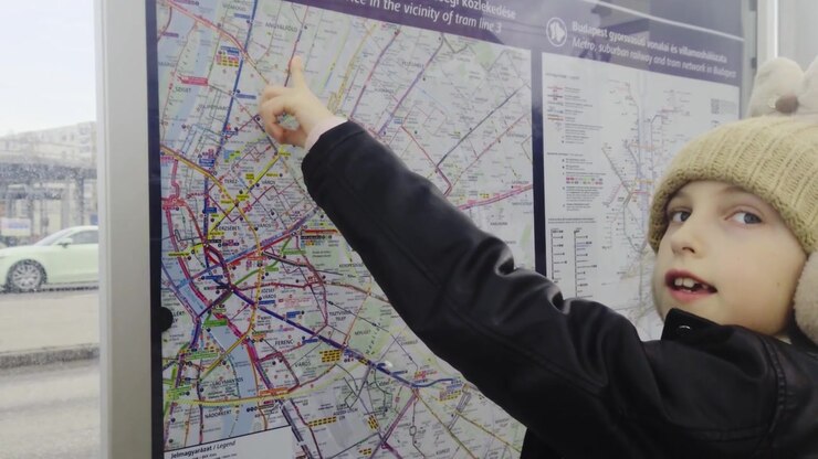 Outdoor medium shot captures a young girl wearing a winter hat and black jacket pointing to a Budapest public transit map inside a street shelter while blurred background traffic moves by.