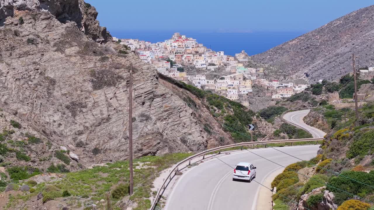 Drone footage follows a car in the foreground, revealing the colorful traditional village of Olympos in Karpathos Island with its stunning hillside and authentic Greek atmosphere