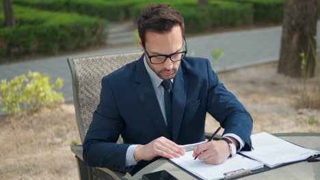 Business-Man-in-Suit-Making-Notes-on-Papers-in-Clipboard-Sitting-by-the-Table-Outdoors---close-up-dolly-in