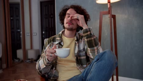 a pensive brunette guy with curly hair drinks tea from a white cup and looks around while thinking about life at home in a modern apartment