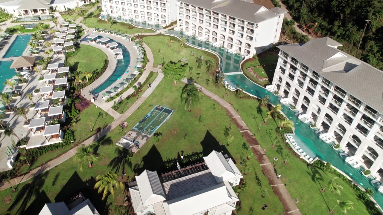 Alejamiento de dron desde suites de hotel de lujo con piscinas de acceso directo, pasando por jardines tropicales cuidados para revelar un grupo de villas boutique y la infraestructura del complejo costero circundante.